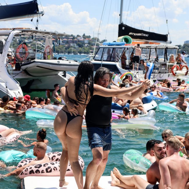 People enjoying a sunny day at a crowded boat party, with various individuals on floaties in the water and a couple dancing on a floating platform. Several boats are anchored nearby as part of a Sydney boat party hire event.
