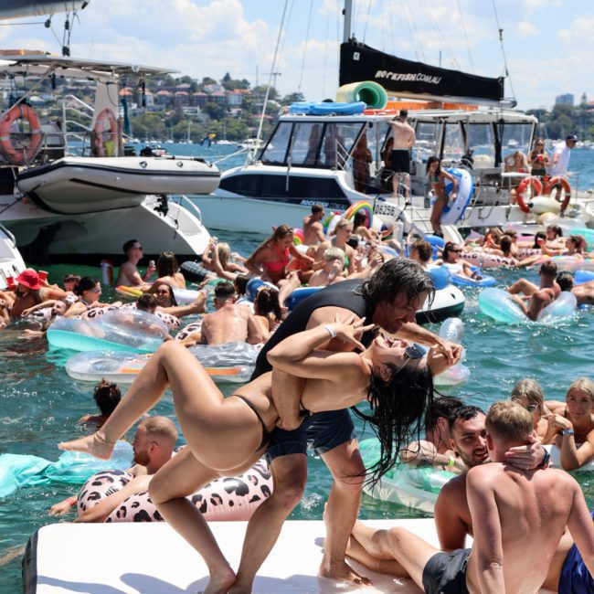 A large group of people enjoying a social gathering on boats and floats in the water on a sunny day. A couple dances on a floating platform while others relax and mingle at a Sydney boat party hire.