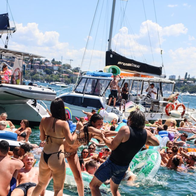 A lively scene of people in swimsuits gathered on boats and inflatables in the water, some dancing and socializing under a clear, sunny sky, enjoying the experience of a Luxury yacht hire Sydney.