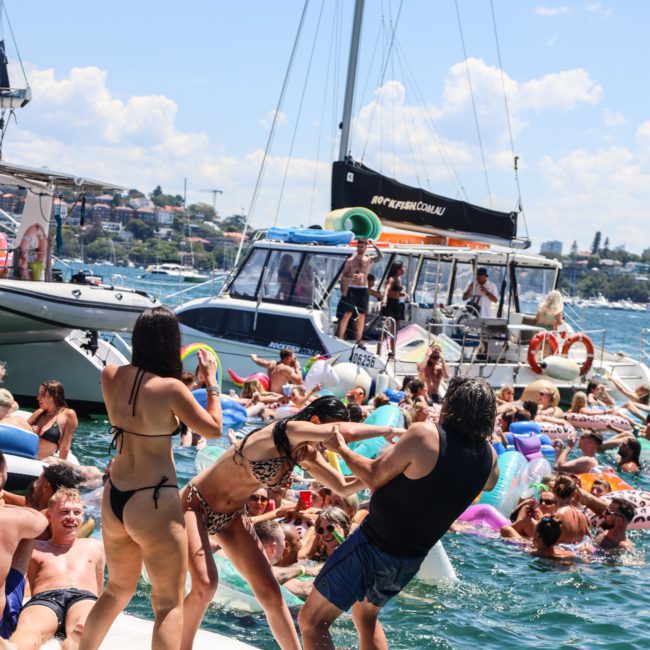 People enjoying a catamaran party Sydney on water with inflatables. Several boats are docked closely together, and attendees are wearing swimwear and engaging in festive activities under a sunny sky.
