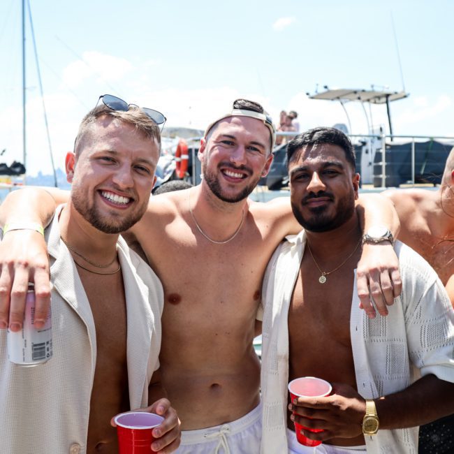 Three men in swimwear pose together on a boat under a sunny sky. Two hold red cups and one holds a beverage can. Boats and water can be seen in the background, making it an ideal scene for a Catamaran party Sydney adventure.