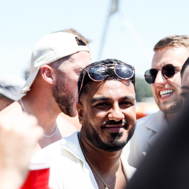 A cheerful group of people outdoors, some wearing sunglasses and hats, smiling and holding red cups. The "Yacht Social Club" logo is visible at the bottom right corner. Experience this vibrant scene with a DJ boat hire Sydney for your next private yacht charter on Sydney Harbour!