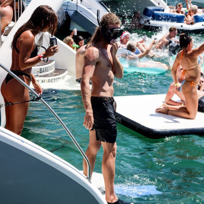 A man wearing a snorkel and flippers stands on a boat's edge, preparing to enter the water. Beside him, a woman uses a phone. Other people swim and lounge on boats and inflatable platforms nearby during a luxury yacht hire in Sydney.