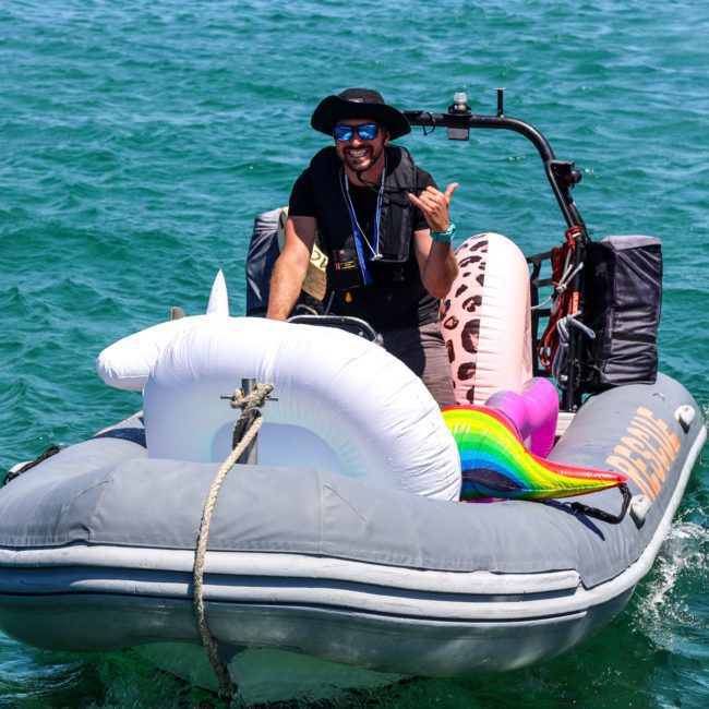 A man in a black outfit and hat steers a small inflatable motorboat on open water, transporting several inflatable pool toys, including a unicorn and a rainbow. Perfect for those considering DJ boat hire Sydney or planning corporate boat events Sydney.