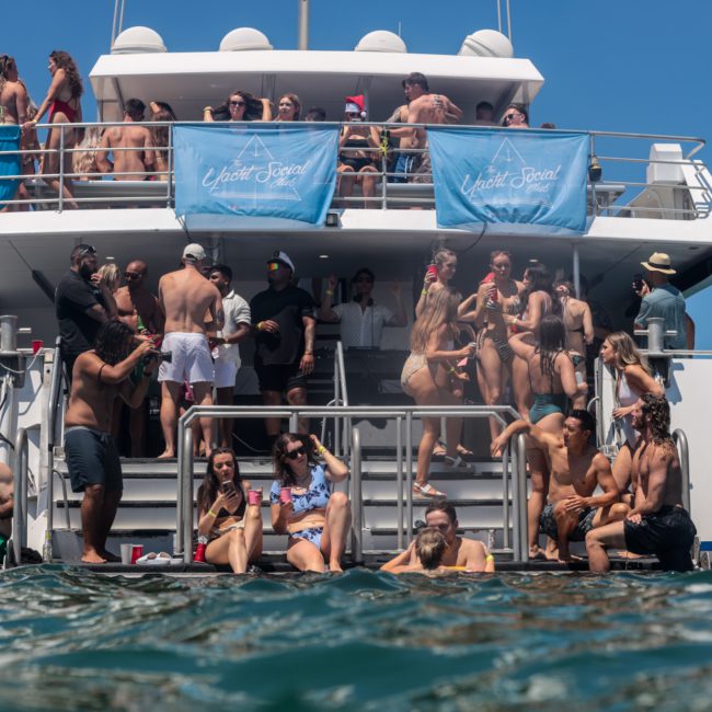 A group of people enjoying a sunny day on a large yacht; some are standing, sitting, or lounging, with banners reading “Yacht Social Club” displayed prominently. Ideal for those interested in Sydney boat party hire or planning corporate boat events in Sydney.
