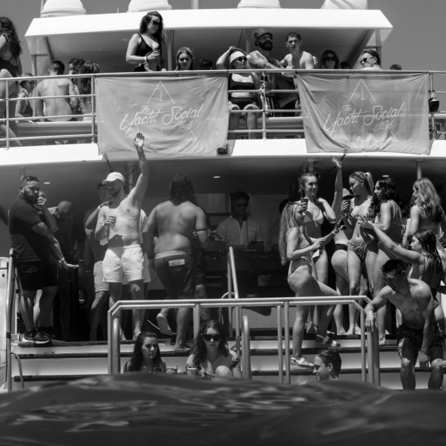 A group of people in swimwear socializing on a multi-deck yacht with "Yacht Social Club" banners displayed, enjoying the Luxury yacht hire Sydney experience.