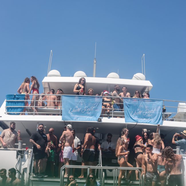 A group of people are gathered on a multi-level boat under a clear blue sky. Some are on the deck, others on the stairs, with blue banners displayed. Many appear to be engaged in social activities. This looks like an ideal event for anyone considering a luxury yacht hire in Sydney.