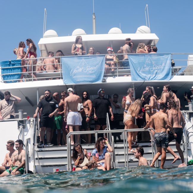 A large crowd of people is gathered on a multi-level boat with "Yacht Social" banners, enjoying a party under the clear blue sky. Some are in swimwear, with water in the foreground. This lively scene captures the essence of a Sydney boat party hire, perfect for those seeking a private yacht charter on Sydney Harbour.