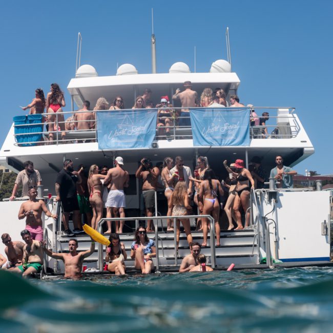 A large group of people are enjoying a sunny day on a double-decker boat. Some are on the lower level near the water, while others are on the upper deck. Banners read "Yacht Social Club." Perfect for corporate boat events Sydney or private yacht charter Sydney Harbour, this scene captures pure joy and leisure.