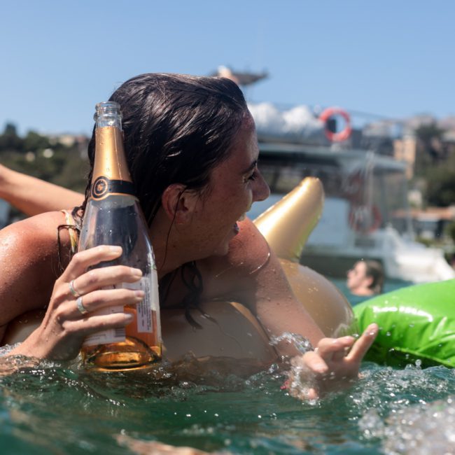 A person is lying on an inflatable float in the water, holding a bottle and smiling with a luxurious yacht in the background. The stunning skyline of Sydney Harbour adds to the serene ambiance, making it an ideal scene for a private yacht charter.