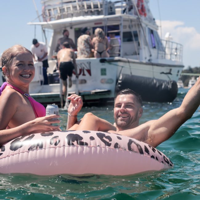 Two people in swimwear float on a leopard print inflatable in the water. The man gives a thumbs-up while others socialize on nearby boats and private yacht charters in Sydney Harbour.