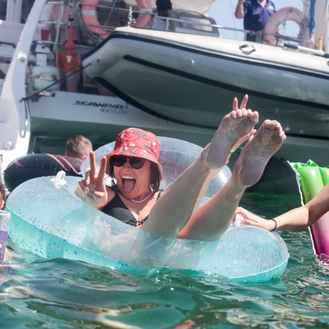 Three people smiling and enjoying their time in the water near boats. One person in a red hat sits in an inflatable ring, playfully posing with their feet up during a lively Sydney boat party hire.