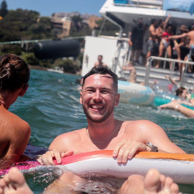 A man floats on an inflatable device in the water, smiling, with a woman nearby. In the background, people enjoy themselves on a private yacht charter Sydney Harbour.