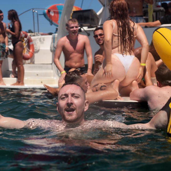 A group of people are enjoying a sunny day on a boat. The man in the foreground is in the water holding a drink and a yellow inflatable. Others are on the boat and in the water, socializing at what appears to be an exciting catamaran party Sydney is known for.