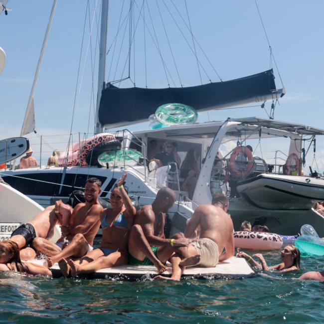 A group of people are lounging on inflatable rafts and floating platforms in the water near several anchored boats on a sunny day, enjoying a Sydney boat party hire.