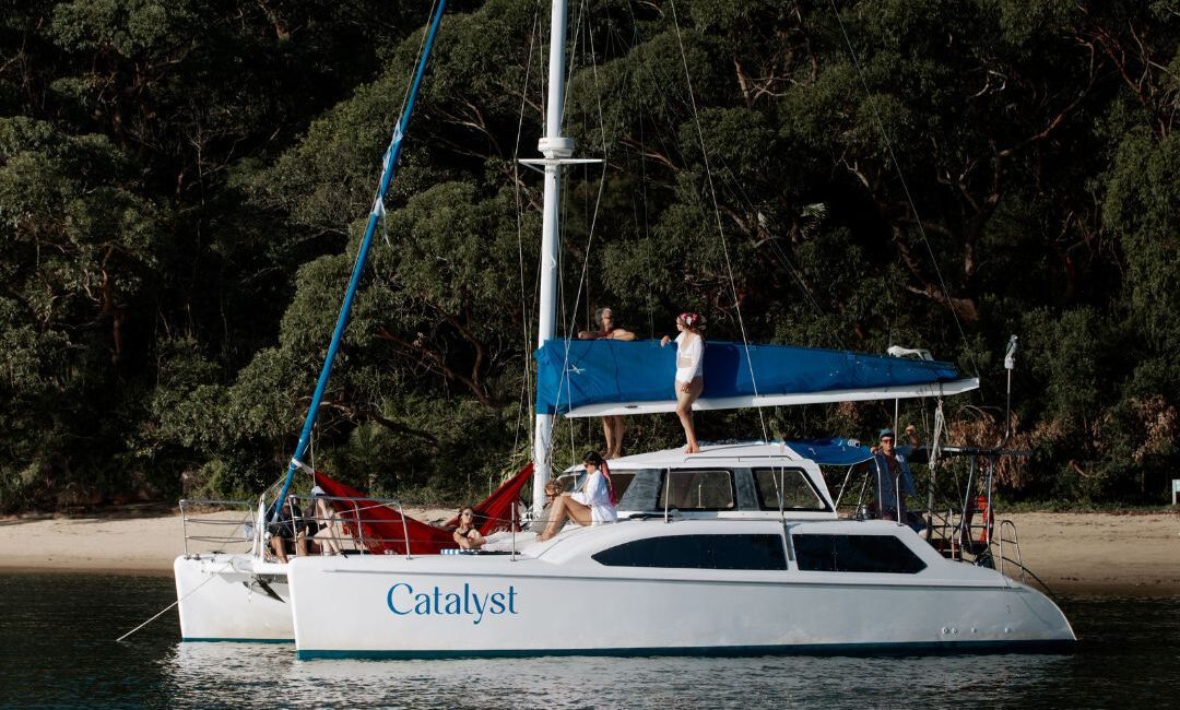 The white catamaran Catalyst is anchored by a forested shore. People are relaxing on deck, with one standing atop the boat and others lounging in red hammocks at the back.