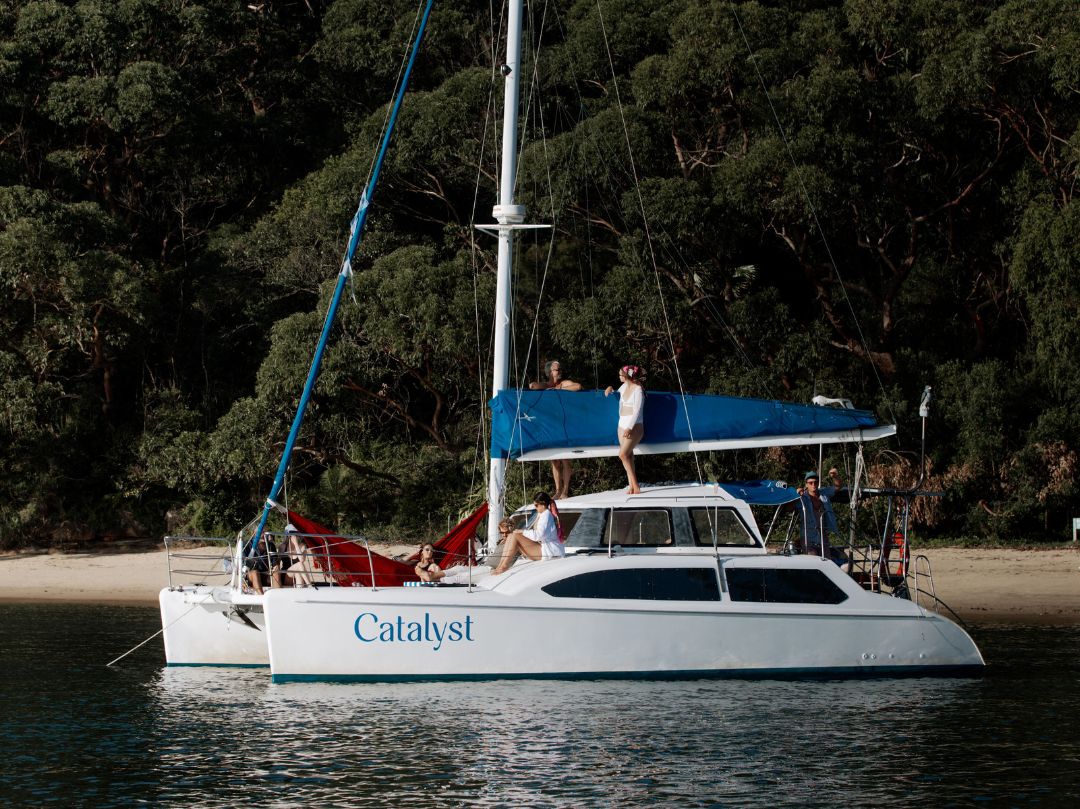 The white catamaran Catalyst is anchored by a forested shore. People are relaxing on deck, with one standing atop the boat and others lounging in red hammocks at the back.
