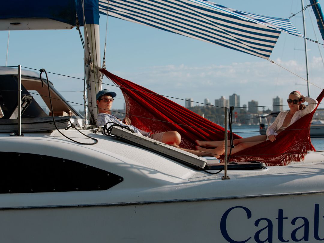 Two people relax on a red hammock under a striped canopy on the sailboat Catalyst, with city buildings and blue sky in the background.
