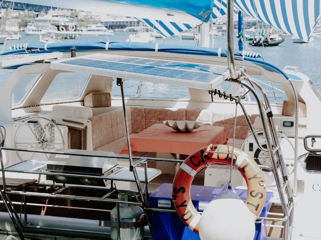 The Catalyst yacht’s deck features cushioned seating, a table with a decorative bowl, striped canopy, solar panel, and lifebuoy. Docked in a marina, Catalyst sits among other boats in the background.