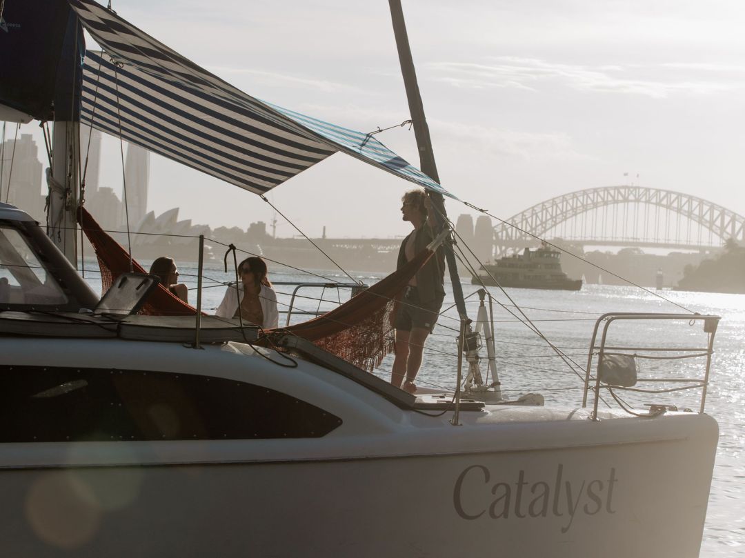Three people relax aboard Catalyst with the Sydney Opera House and Harbour Bridge in the sunny background.