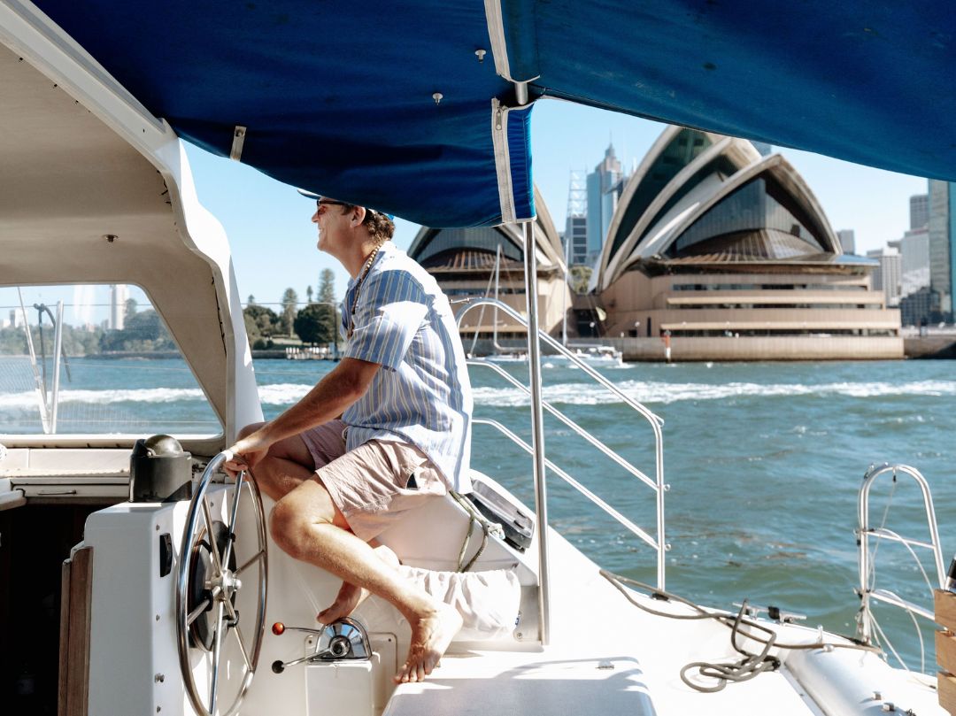 A person steers the Catalyst boat on the water near the Sydney Opera House under a blue canopy, with city buildings visible in the sunny background.