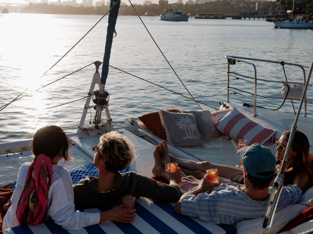 Four people relax on Catalyst at sunset, seated on cushions and holding drinks. Calm water and other boats in the background create a peaceful, leisurely atmosphere.