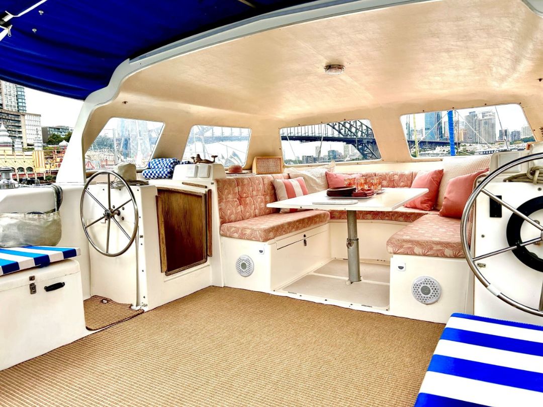 Catalyst boat interior features U-shaped cushioned seating, pink patterned pillows, a white table with drinks, dual steering wheels, striped blue and white cushions, and large windows with city and bridge views.