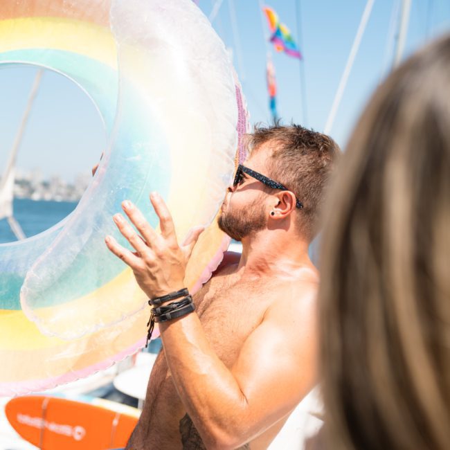 A shirtless man wearing sunglasses holds a large inflatable ring with rainbow colors on a luxury yacht under clear blue skies.