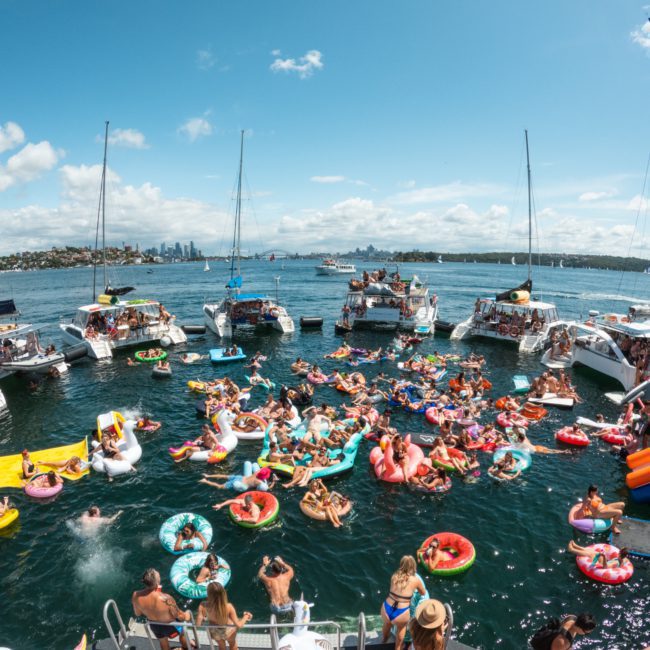 People relax and socialize on inflatable floats and boats gathered on a sunny day in a body of water with a city skyline in the background, reminiscent of a vibrant catamaran party in Sydney.