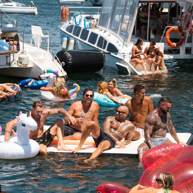 People relaxing on flotation devices and swimming while gathered around boats on a sunny day at the lake, with a boat named "Rum Runner Cove" in the background. This scene is perfect for those considering a Sydney boat party hire or corporate boat events in Sydney to enjoy a day out on the water.