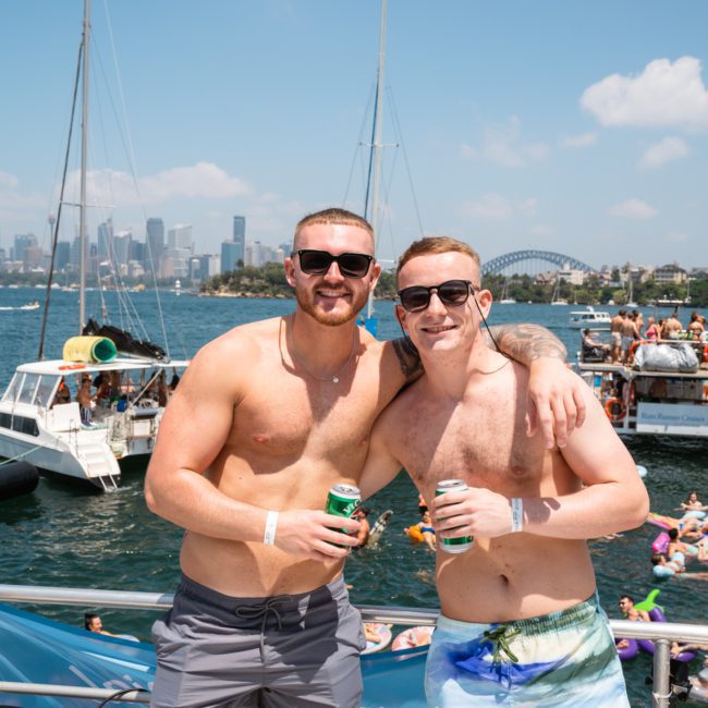 Two men in swim shorts and sunglasses pose with drinks on a boat. Other boats and people swimming are visible in the background. The Sydney Opera House and Harbour Bridge can be seen in the distance, making it a perfect setting for corporate boat events Sydney.