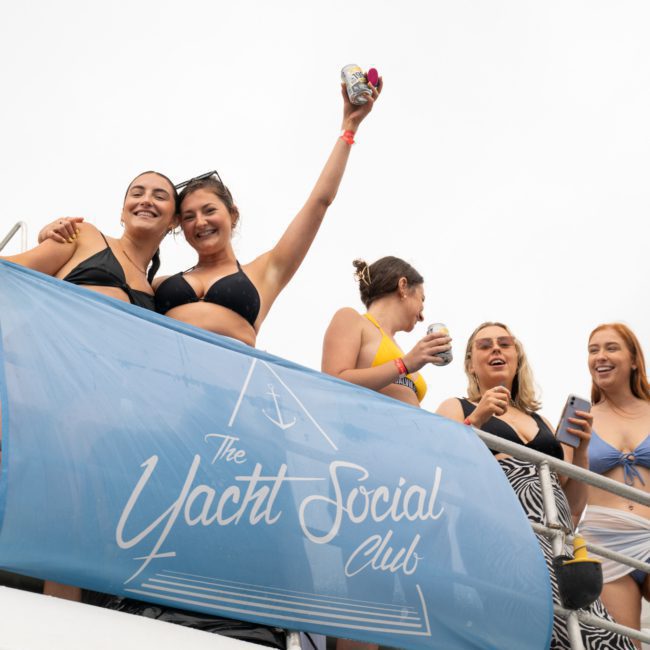 Five people in swimwear enjoy drinks on a yacht for a Sydney boat party hire. A banner reads "The Yacht Social Club.