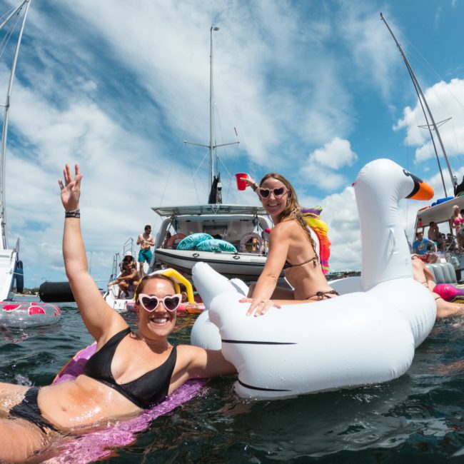 Two people are enjoying the water on inflatable floats near anchored boats under a partly cloudy sky. One is on a swan-shaped float, and another waves while lying on a yellow float, relishing in the ambiance that only a catamaran party Sydney can offer.