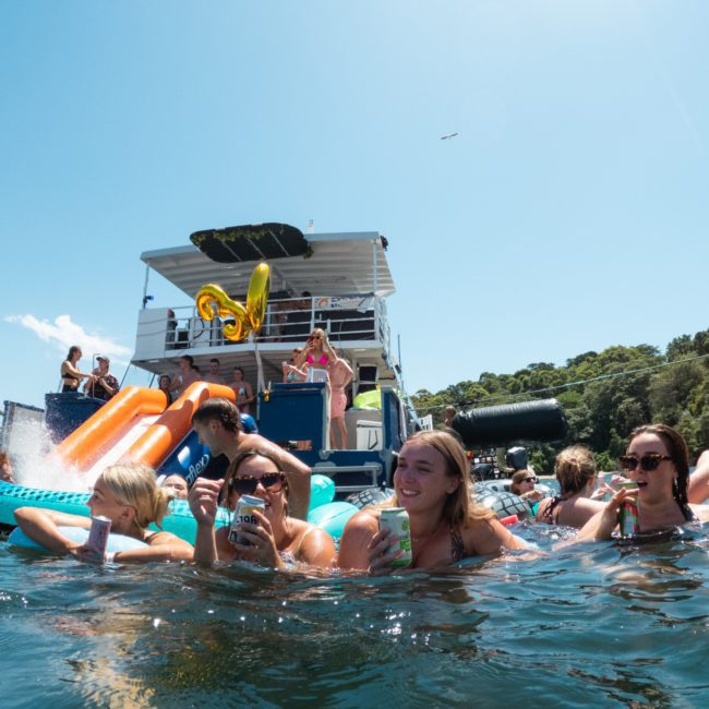 A group of people enjoys swimming and lounging on floaties in a body of water near a boat, under a sunny sky, making the most of their Sydney boat party hire.