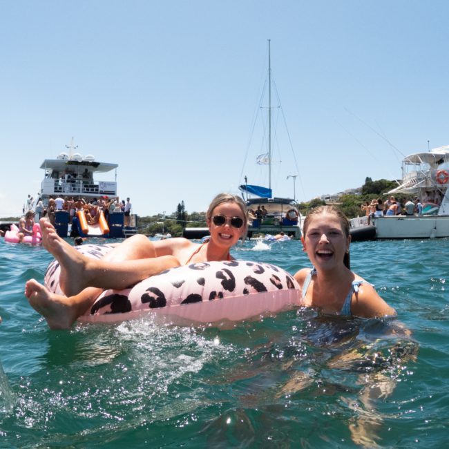 Two women enjoy a sunny day in the water, one on a flamingo float and the other swimming, with boats and people in the background, perfect for a Sydney boat party hire.
