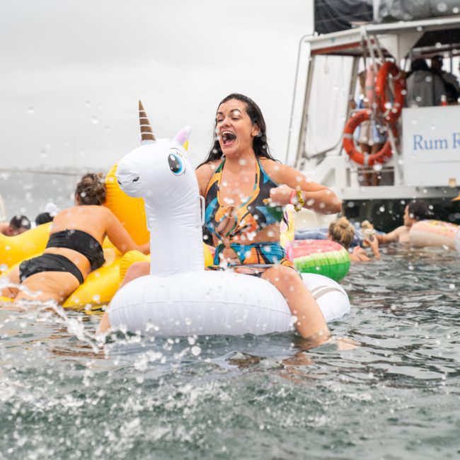 A woman wearing a swimsuit rides an inflatable unicorn while splashing in water among other people and inflatables near a boat labeled "Rum Runner Cruises," enjoying a lively Catamaran party Sydney.