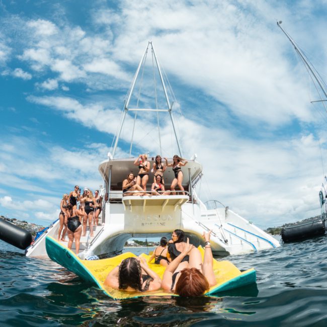 Group of people on a boat, with some lounging on a floating mat in the water. Clear blue sky and another boat visible in the background. Perfect day for a Catamaran party Sydney-style!