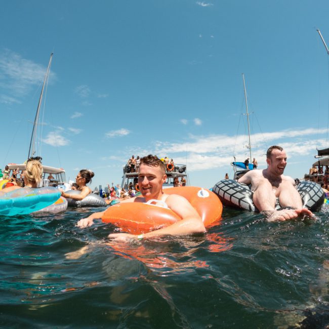 People floating on inflatable rafts in the water near boats under a clear blue sky, enjoying their time during a catamaran party Sydney.