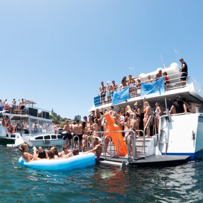 A group of people socializing on a docked luxury yacht hire Sydney while others relax in an inflatable blue pool in the water on a sunny day. Other boats are visible in the background.