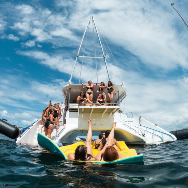 People enjoying a sunny day on a white boat, with some swimming in the water on yellow and green inflatables. The sky is blue with scattered clouds, perfect for a catamaran party in Sydney.