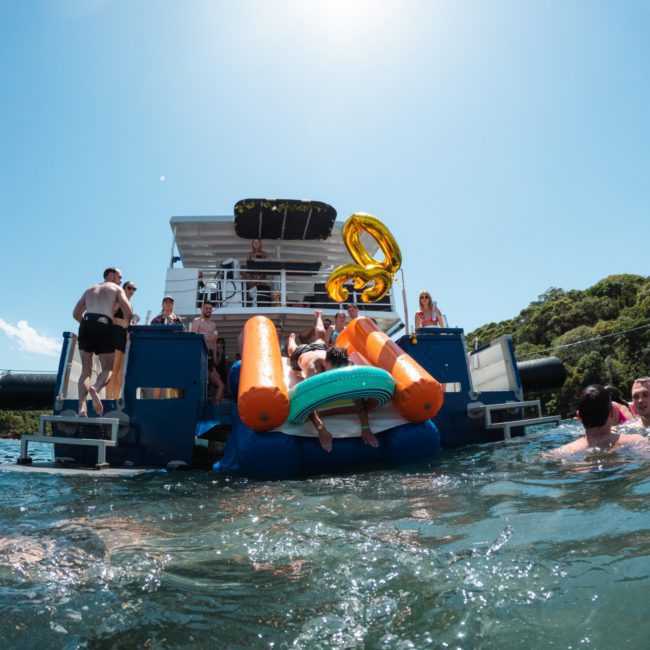People enjoying a sunny day on a private yacht charter in Sydney Harbour with large inflatables in the water. Some individuals are climbing back onto the boat, while others swim nearby. The boat is anchored near a lush, green coastline.