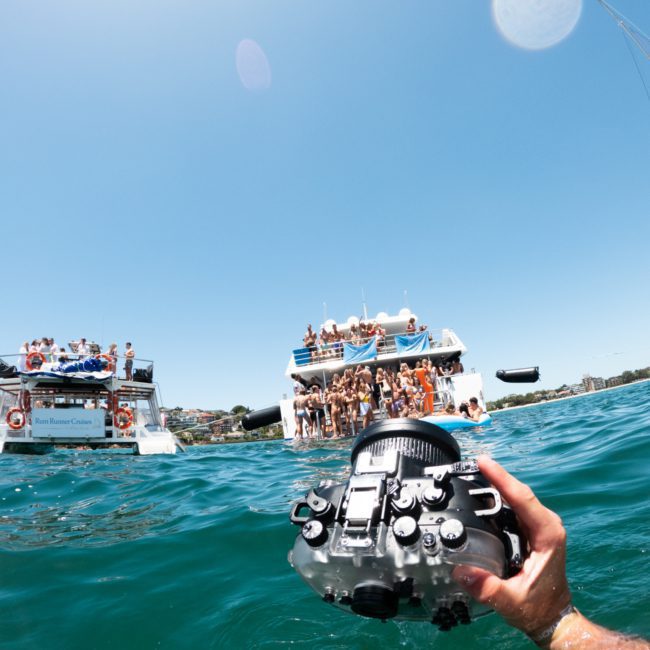 A person's hand holds an underwater camera partially submerged in water, with two boats in the background filled with people enjoying a sunny day at a Sydney boat party hire.
