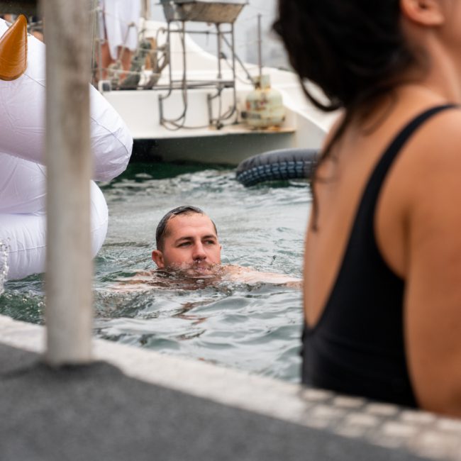 A person swims in a pool near an inflatable unicorn while others enjoy the surroundings. In the foreground, someone in a low-cut black top partially obstructs the view. Consider enhancing your day with a private yacht charter on Sydney Harbour for an unforgettable experience.
