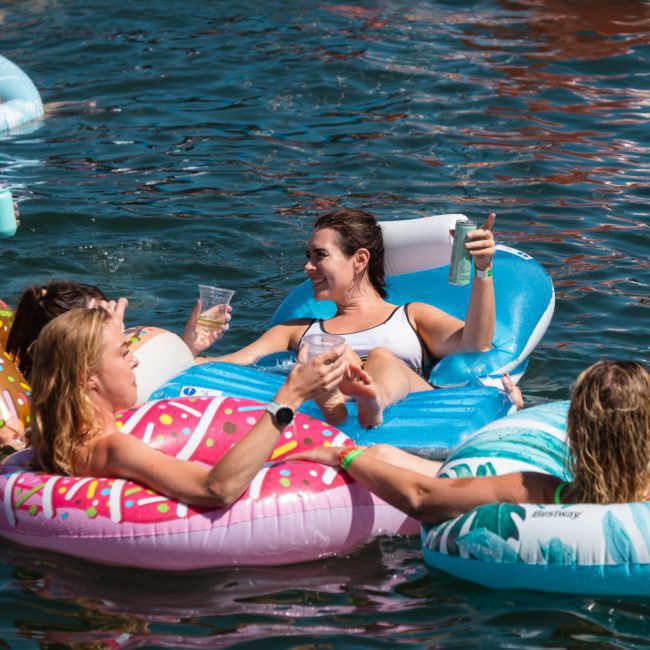 People are relaxing on colorful inflatable pool floats in the water, holding drinks and enjoying a sunny day during a catamaran party in Sydney.