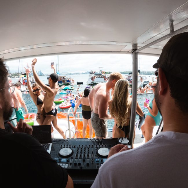 A DJ duo performs on a boat while a large crowd in swimwear dances and socializes, with inflatable toys visible. The backdrop includes city skyline and watercraft. Perfect for those seeking luxury yacht hire Sydney or planning corporate boat events Sydney.