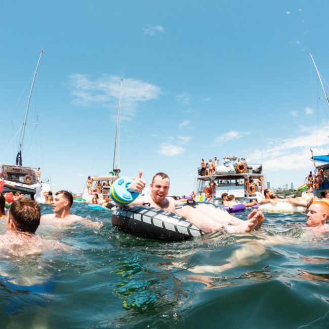 People are enjoying a sunny day in the water with various flotation devices and boats, including a luxury yacht hire Sydney, in the background.