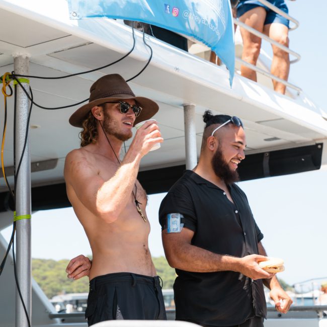 Two people stand on a boat under a "Yacht Social Club" banner. One person is shirtless wearing a hat and sunglasses, the other is wearing a black shirt and holding a donut-like item. Perfect setup for your next DJ boat hire Sydney experience!