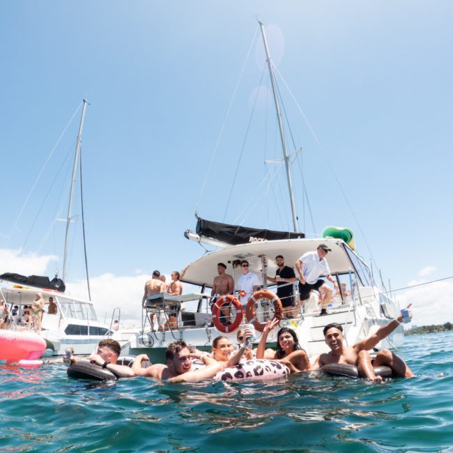 People enjoying time in the water near a private yacht charter Sydney Harbour under a clear sky. Some are floating on inflatables while others are on the boat.