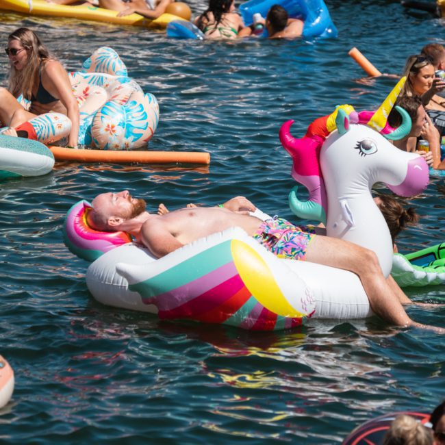 A man is relaxing on a colorful unicorn float in the water, surrounded by other people on various inflatables near a corporate boat event in Sydney.