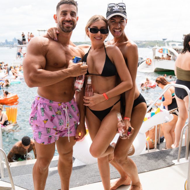 Three people in swimwear pose smiling with drinks on a catamaran during a sunny day, while others swim and float in the water in the background, enjoying a Sydney boat party hire.